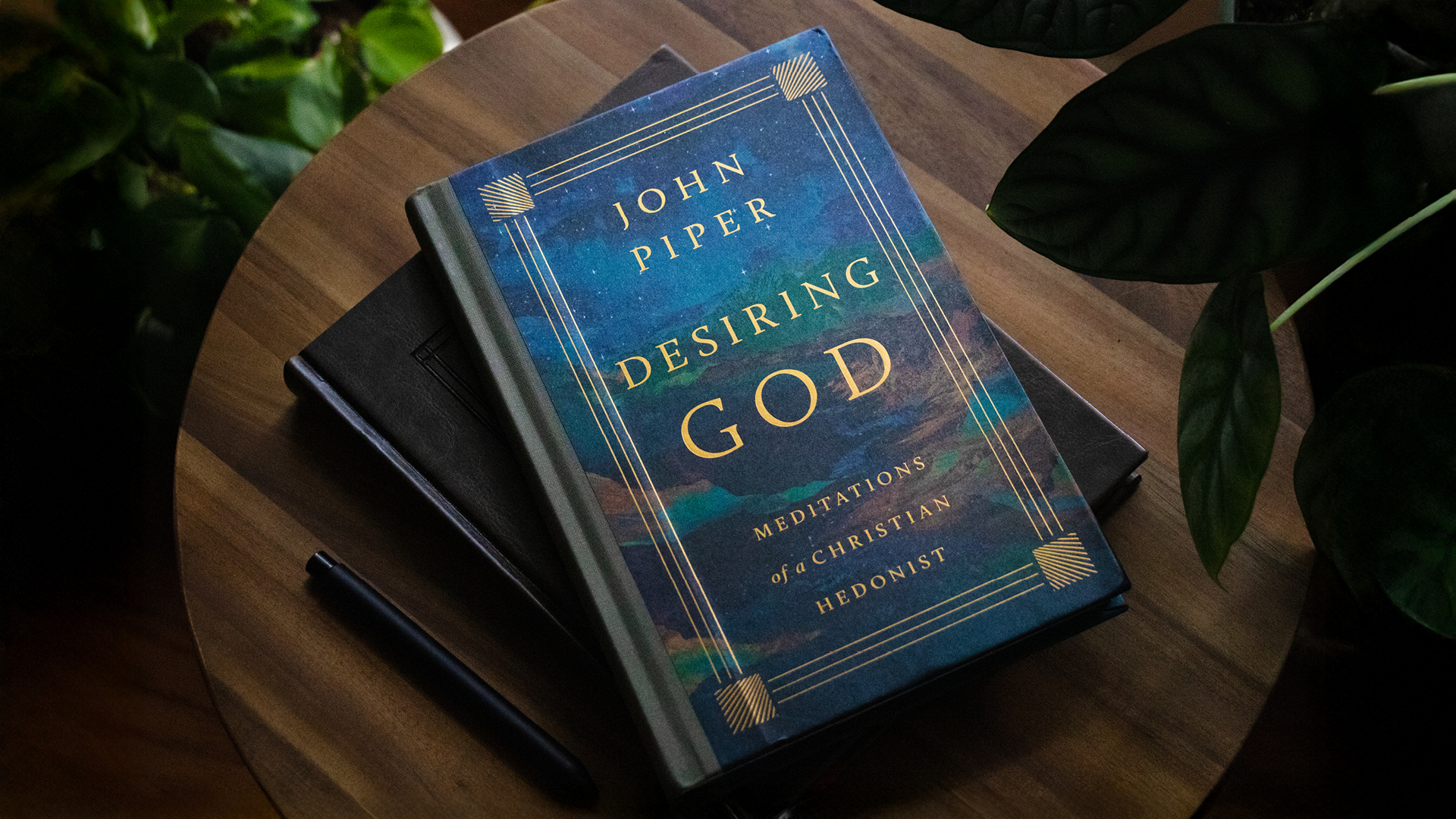 A copy of John Piper's book 'Desiring God' on the top of a pile of books on a wooden table.